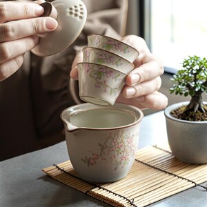 May include: A beige ceramic tea set with floral designs. The set includes a teapot with a lid, and three small teacups. The teacups and teapot are decorated with pink and green floral patterns. The tea set is on a bamboo mat.