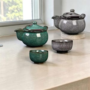 Peut inclure: Un service à thé en céramique vert et gris, comprenant deux théières et deux petites tasses. Les théières ont des couvercles et des becs verseurs. Une théière et une tasse sont vertes, l'autre théière est grise. L'ensemble est présenté sur une surface claire.