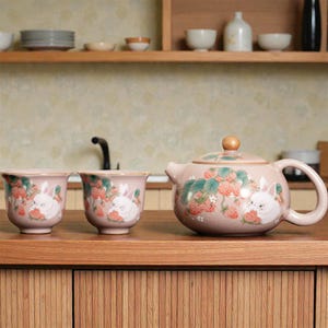 May include: A pink ceramic tea set featuring a teapot and two cups. The set is decorated with a strawberry and bunny design. The teapot has a wooden knob on the lid and a curved handle. The cups have a rounded shape.