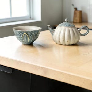 Pode incluir: Um conjunto de chá de cerâmica com uma tigela azul-petróleo com um design floral e uma chaleira branca em forma de abóbora com tampa azul. O conjunto está sobre uma superfície de madeira clara, adequado para cozinha ou sala de jantar.