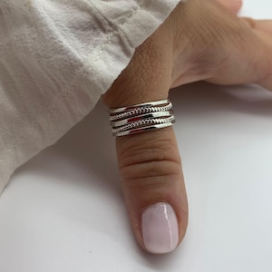 May include: A silver ring with three bands, each featuring a different design. The top and bottom bands have a smooth, polished finish, while the middle band has a textured, beaded pattern. The ring is worn on a finger.