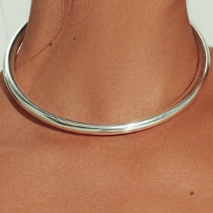 May include: A smooth, polished silver-toned choker necklace featuring a thick, rounded, continuous band design that sits closely around the neck. The metal surface has a reflective, high-shine finish that catches the light evenly across its circular form.