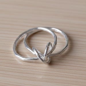 May include: A silver-colored ring with a knot design. The ring features two intertwined bands that form a knot in the center. The ring is made of a shiny, metallic material and is set against a light brown wooden surface.