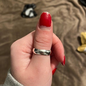 Può includere: Un anello argentato lucido con un design liscio e arrotondato. L'anello è indossato su un pollice con smalto rosso. L'anello riflette l'ambiente circostante.