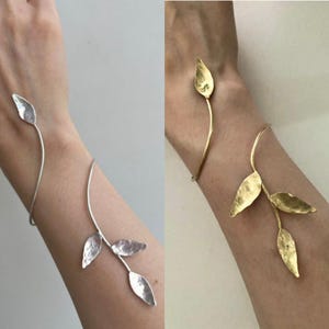 Può includere: Un bracciale a fascia in metallo color argento caratterizzato da un design a stelo sottile e curvo con tre foglie martellate. Il bracciale avvolge il polso, mettendo in mostra una finitura metallica testurizzata sulle foglie e una struttura sottile.