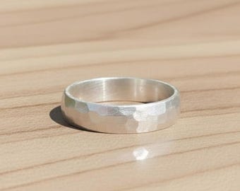 May include: A silver ring with a textured, faceted surface. The ring is round and has a wide band. The ring is set against a light brown wooden surface.