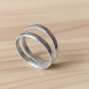 Può includere: Un anello d'argento a doppia fascia. L'anello presenta due fasce parallele collegate alla base. Le fasce hanno una finitura liscia e lucida. L'anello è appoggiato su una superficie di legno marrone chiaro.