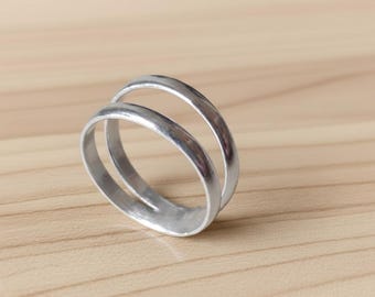 Könnte beinhalten: Ein silberner Doppelring. Der Ring hat zwei parallele Bänder, die an der Basis verbunden sind. Die Bänder haben eine glatte, polierte Oberfläche. Der Ring liegt auf einer hellbraunen Holzoberfläche.