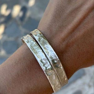 May include: Two silver-colored bangle bracelets with a hammered, textured surface. The bracelets have a reflective, metallic finish and a rounded, solid band construction. They are worn together on the wrist, showcasing the uneven, dimpled pattern.