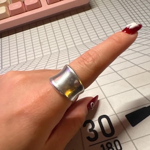 May include: A silver ring with a curved, wide band. The ring is smooth and reflective, catching the light. The ring is worn on a finger.