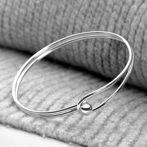 May include: A polished silver-tone bangle bracelet featuring a sleek, minimalist double-wire design that converges at a single, smooth spherical bead. The metallic surface reflects light, highlighting the continuous, rounded form of the jewelry piece.