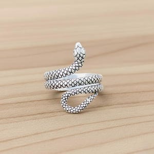May include: A silver-coloured ring in the shape of a snake. The snake's body coils around the finger, with the head raised. The ring has a textured, scale-like pattern. The ring is on a light brown wooden surface.