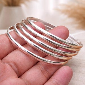 May include: A stack of six shiny, silver bangles. The bracelets are smooth and round, reflecting light. The jewelry is held in a hand, with a soft, neutral background.