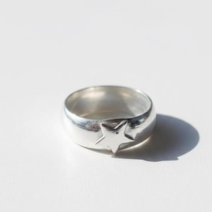 May include: A silver ring featuring a star motif. The band ring has a raised star design. The ring is made of a shiny, reflective metal. The ring is displayed on a white surface.