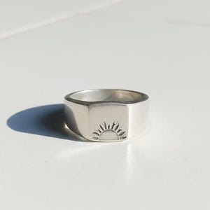 Könnte beinhalten: Ein silberner Siegelring mit rechteckiger Oberfläche. Der Ring hat ein eingraviertes Sonnenmuster auf der Oberfläche. Der Ring besteht aus einem glänzenden, metallischen Material.
