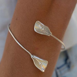 May include: A silver-toned open cuff bracelet featuring two stylised calla lily flower motifs at each end. The metal petals have a smooth, reflective surface with small, raised spherical details at the centre of each flower head, connected by a thin, flexible band.