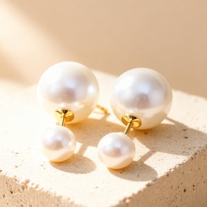 May include: A pair of elegant pearl earrings. Each earring features a large, lustrous white pearl and a smaller pearl, connected by a gold-colored post. The earrings are set against a neutral background, highlighting their classic design.