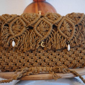 May include: A brown macrame handbag with a rectangular shape and a flap closure. The bag features a woven design with fringe detailing and a braided strap. It is adorned with small gold charms and white teardrop-shaped beads.