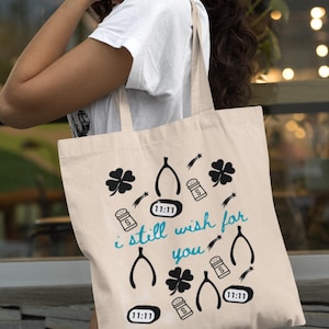 Könnte beinhalten: Beige Tragetasche mit schwarzen und türkisfarbenen Grafiken. Das Design umfasst vierblättrige Kleeblätter, Wunschknochen, Salzstreuer, Sternschnuppen und die Zahl 11:11. Der türkisfarbene Text lautet "i still wish for you".