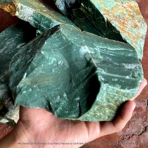 May include: A large, raw chunk of deep green stone with a smooth, crystalline texture and jagged, uneven edges. The mineral displays variations in shade from dark forest green to lighter translucent tones, featuring natural fractures and rough surfaces.