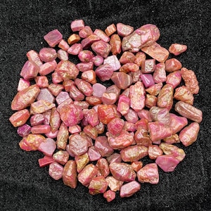 May include: A collection of raw, unpolished ruby gemstones in various shades of pink and deep red. These natural mineral specimens feature irregular, jagged shapes with a mix of translucent and opaque surfaces, showcasing a rough, crystalline texture.