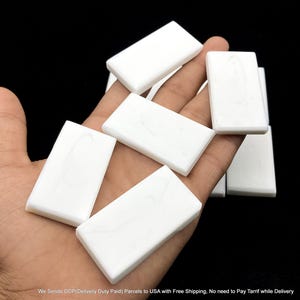 Puede incluir: Una colección de baldosas rectangulares de color blanco sólido con un acabado liso y pulido, que presentan sutiles patrones de veteado gris. Estas piezas planas y uniformes están apiladas, mostrando sus bordes limpios y forma constante.
