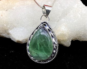 May include: A teardrop-shaped green gemstone pendant set in a polished silver frame with a twisted rope border. The pendant hangs from a thin silver snake chain, showcasing the translucent, multifaceted surface of the stone and its metallic setting.
