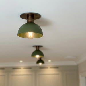 Op de afbeelding: Olijfgroene en goudkleurige plafondlampen in de vorm van een koepel met een messing voet. De lampen zijn in een rij gerangschikt, waarbij de dichtstbijzijnde scherp is. De binnenkant van de koepel is goudkleurig en de gloeilamp is zichtbaar.