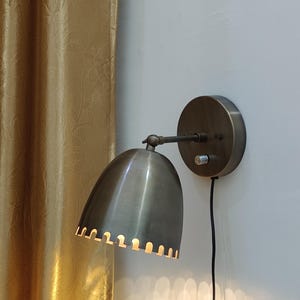 May include: A wall-mounted lamp in an antique style, with a brushed metal finish. It has a rounded shade with a scalloped edge, a pivoting arm, and a circular base. A black cord extends from the lamp. The background includes a gold curtain and a white wall.
