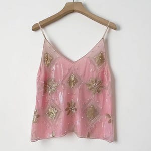 Pode incluir: Um top camisola rosa com decote em V e alças finas, adornado com lantejoulas douradas e padrões florais e de diamantes. O top está pendurado numa cabide de madeira sobre um fundo branco.