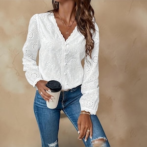 May include: White eyelet blouse with long sleeves and a V-neck. The blouse has a button-down front and floral cut-out details. The model is wearing blue jeans and holding a coffee cup.