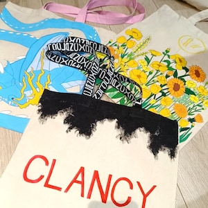 Può includere: Diverse borse tote in tela con vari disegni. Una presenta un drago blu, un'altra fiori gialli, e una terza mostra il nome "CLANCY" in rosso. Le borse hanno cinghie di diversi colori, tra cui rosa e una cinghia con motivo bianco e nero.