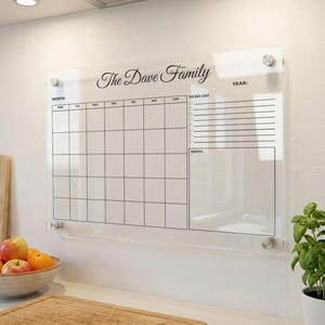 May include: Clear acrylic family calendar with the text "The Dave Family" in elegant script. The calendar includes a monthly grid, a to-do list, and a notes section. It is mounted on a white wall with silver hardware. A bowl of fruit and a plant are in the foreground.