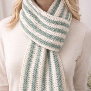 May include: A cream and pale green striped scarf, knitted with a soft material. The scarf is wrapped around the neck, showcasing a simple, elegant design. The scarf's measurements are in centimetres.