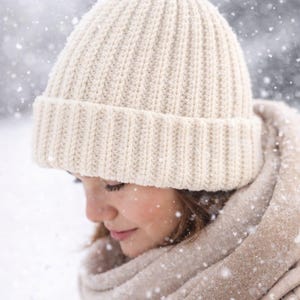 May include: Cream-colored knit beanie with a folded brim. The hat is ribbed and appears warm. A matching scarf is wrapped around the neck. Snowflakes fall in the background, suggesting cold weather. The image focuses on winter apparel.