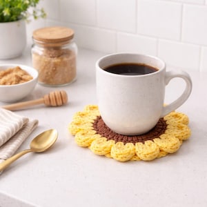 Może przedstawiać: Biały ceramiczny kubek wypełniony ciemną kawą stoi na ręcznie robionym szydełkowym podkładce w kształcie słonecznika. Podkładka ma żółte płatki i brązowy środek. Inne przedmioty to łyżka, nabierak do miodu i cukier.