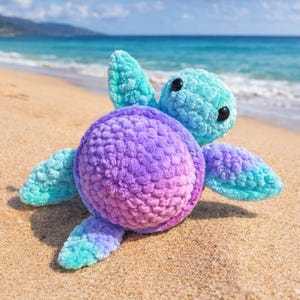 Pode incluir: Uma tartaruga marinha de crochê feita à mão com um corpo redondo em tons de roxo e rosa, e barbatanas e cabeça turquesa. A tartaruga tem olhos pretos e está em uma praia arenosa com o oceano ao fundo.