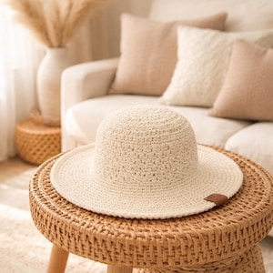 May include: A cream-colored, crocheted sun hat with a wide brim sits on a round, woven side table. The hat has a small, brown leather tag. The table is made of woven wicker and has three wooden legs. The background includes a sofa and decorative pillows.