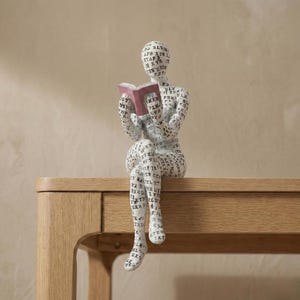 Könnte beinhalten: Eine weiße Figur einer Person, die ein Buch liest. Die Figur ist mit schwarzem Text bedeckt. Das Buch hat einen weinroten Einband. Die Figur sitzt auf einer Holzoberfläche.