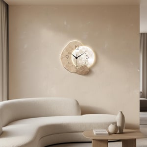 Puede incluir: Un reloj de pared moderno con un diseño abstracto único. La esfera del reloj es de color beige claro con manecillas y números negros. El reloj tiene forma circular con una luz brillante en los bordes. El reloj está montado en una pared beige.