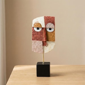 May include: Abstract face sculpture with a geometric design. The sculpture features a colour palette of red, white, and tan. It is mounted on a gold-coloured rod and a black square base. The artwork is a decorative piece.
