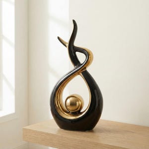 Può includere: Una scultura in ceramica nera e oro con finitura lucida. Il design astratto presenta forme intrecciate e una sfera dorata al centro. La scultura è esposta su una superficie in legno di colore chiaro.