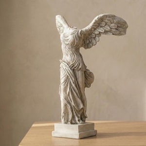 Puede incluir: Estatua detallada en blanco roto de la Victoria de Samotracia. La escultura representa una figura femenina drapeada sin cabeza con grandes alas detalladas. La estatua está montada sobre una base rectangular y sobre una superficie de madera.