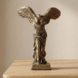 Puede incluir: Estatua de bronce de la Victoria de Samotracia, una escultura clásica. La estatua representa una figura alada drapeada, con alas extendidas, de pie sobre una base rectangular. La escultura tiene una apariencia envejecida y antigua.