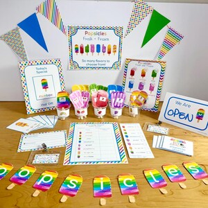 May include: A popsicle stand play set featuring colorful signs with text like Popsicles Fresh + Frozen, Today's Special, and We Are OPEN. Includes a banner spelling popsicles, flavor cards, a tracker, and small plastic cups with popsicle cutouts.