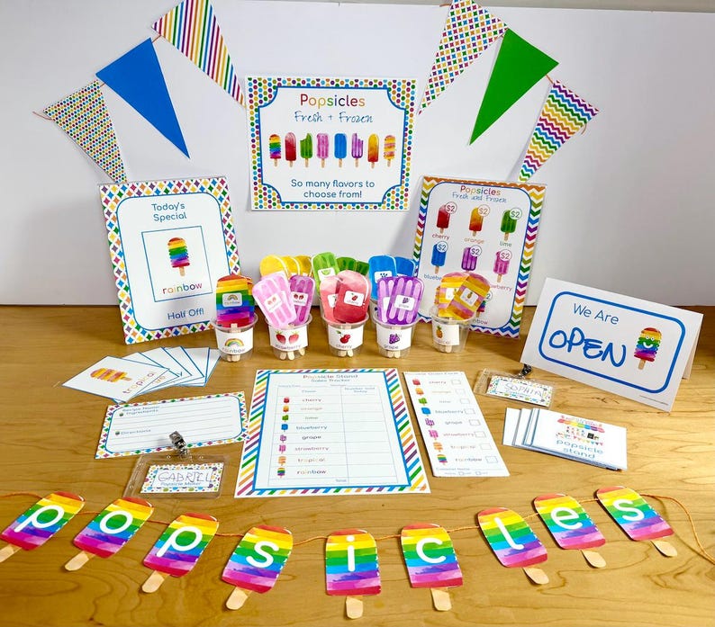 May include: A popsicle stand play set featuring colorful signs with text like Popsicles Fresh + Frozen, Today's Special, and We Are OPEN. Includes a banner spelling popsicles, flavor cards, a tracker, and small plastic cups with popsicle cutouts.