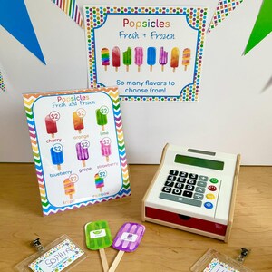 May include: A popsicle shop play set featuring a sign with the text Popsicles Fresh + Frozen, a menu board with various popsicle flavors and prices, a white toy cash register, two popsicle cutouts, and two name badges labeled Sophia and Lukas.
