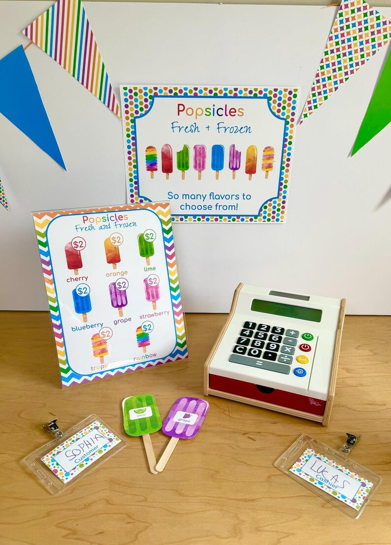 May include: A popsicle shop play set featuring a sign with the text Popsicles Fresh + Frozen, a menu board with various popsicle flavors and prices, a white toy cash register, two popsicle cutouts, and two name badges labeled Sophia and Lukas.