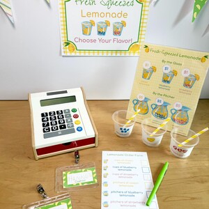 May include: A lemonade stand set featuring a white toy cash register, three clear plastic cups with yellow striped straws, a printed menu sign, a lemonade order form with a green pen, and two name badges labeled Hannah and Mateo.