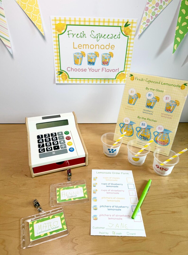 May include: A lemonade stand set featuring a white toy cash register, three clear plastic cups with yellow striped straws, a printed menu sign, a lemonade order form with a green pen, and two name badges labeled Hannah and Mateo.